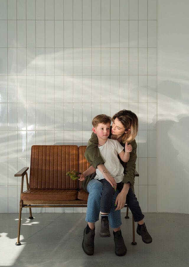 Recto de la carte : une mère serre son petit garçon contre elle, assis sur un banc dans une pièce baignée de lumière