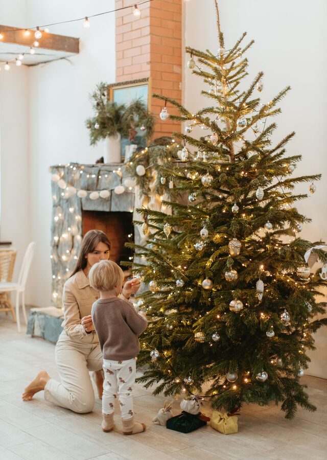 Recto de la carte : une mère et son petit garçon accrochent les boules du sapin, près de la cheminée